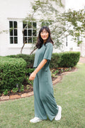 Female model standing outdoors in a garden wearing the Women's Wide Leg Pant in Pine and matching short sleeve tee on white shoes