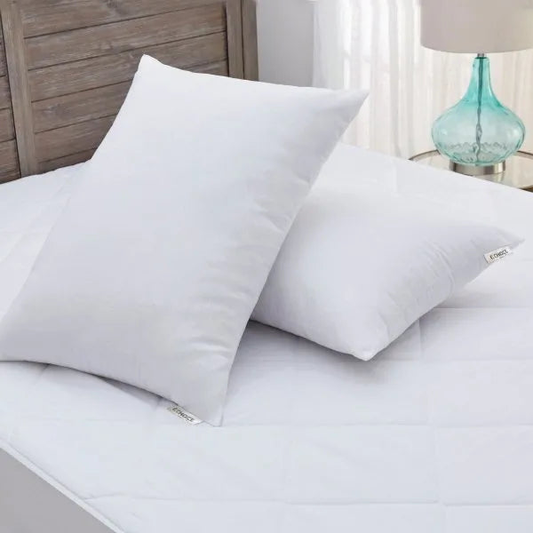 Two white Keeco Woodspring Ecoendure Pillows, as featured at many Choice® Hotels, rest on a quilted mattress—one upright by a wooden headboard, bathed in sunlight through sheer curtains and beside a blue glass lamp. Ideal for stomach sleepers.