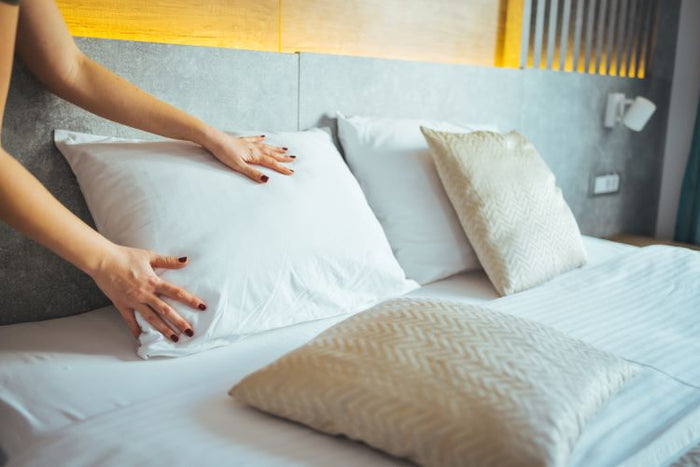 A person with dark red nails arranges a Keeco Woodspring Ecoendure Pillow, featured at many Choice® Hotels, offering medium-firm support on a neatly made bed with light decorative pillows, a modern gray headboard, and warm lighting.