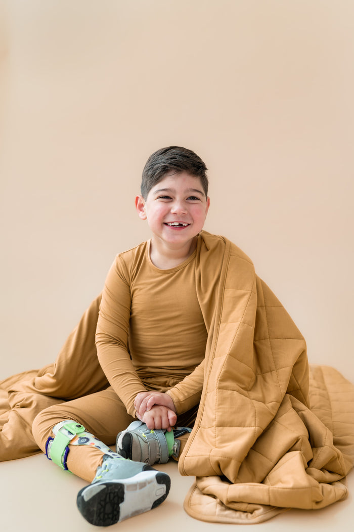 Child covered with Youth Blanket in Ochre 2.5 wearing a similar color long sleeve pajama set on a beige background