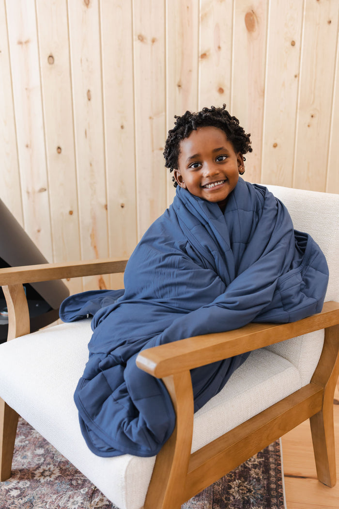 Boy smiling with Youth Blanket in Oxford 2.5 wrapped around him sitting on a white sofa in a room