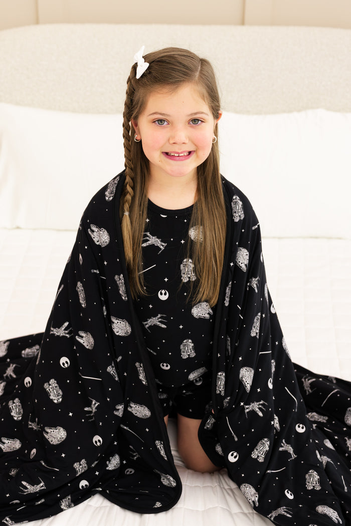 Girl sitting on bed with Youth Blanket in STAR WARS™ 2.5 print wearing similar print short sleeve pajama set
