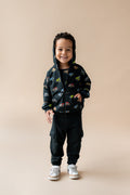 Boy standing wearing Black Zip-Up Hoodie in Monster Truck print over black pant and white shoes on a beige background