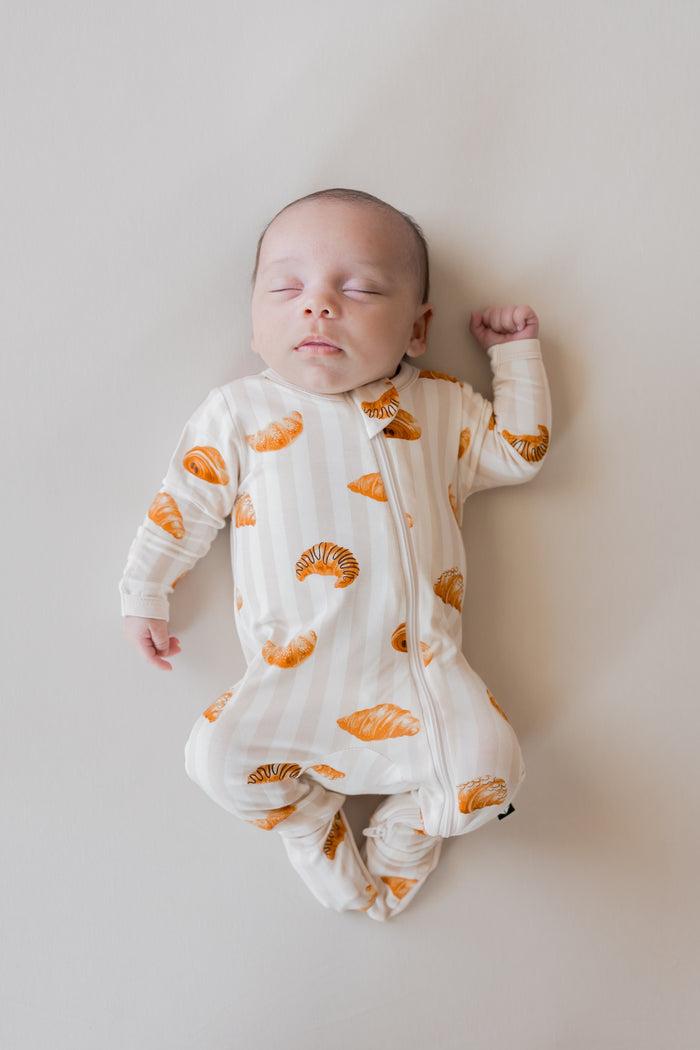 Baby sleeping in Zippered Footie with Croissant print on a beige background