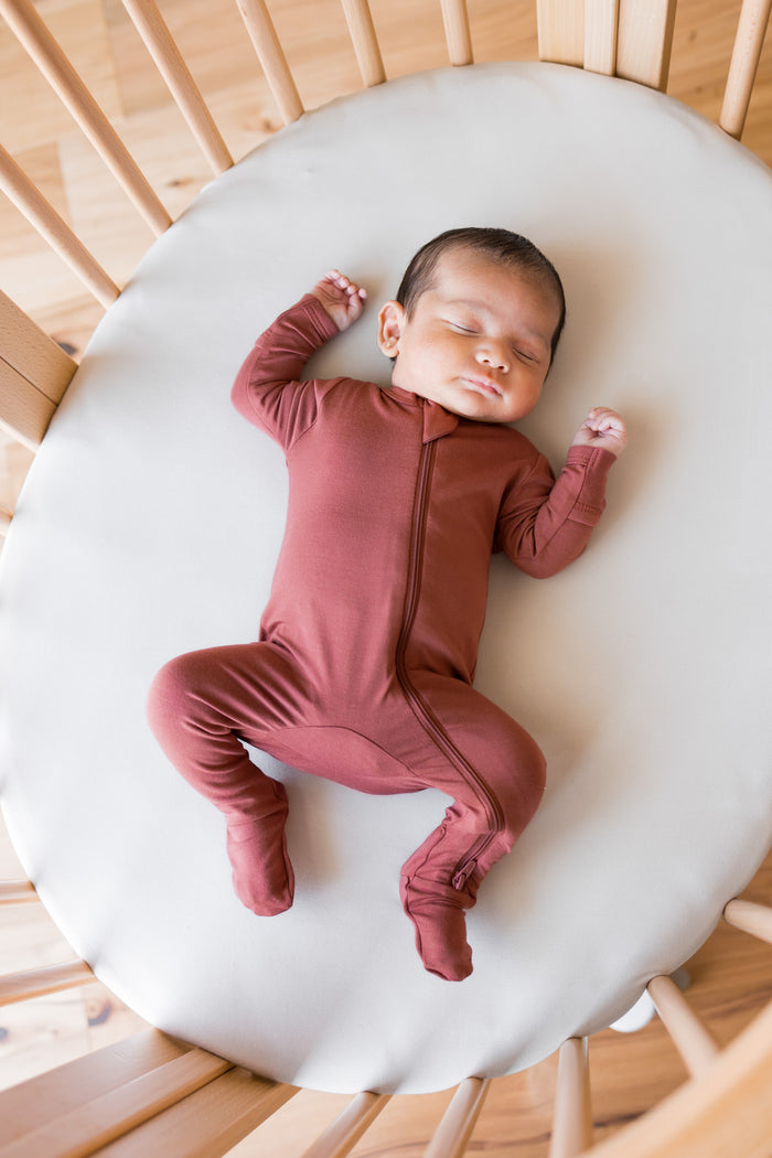 A sleeping newborn baby wearing a dusty terracotta footed zip-up sleeper, lying on a white oval mattress inside a wooden crib.