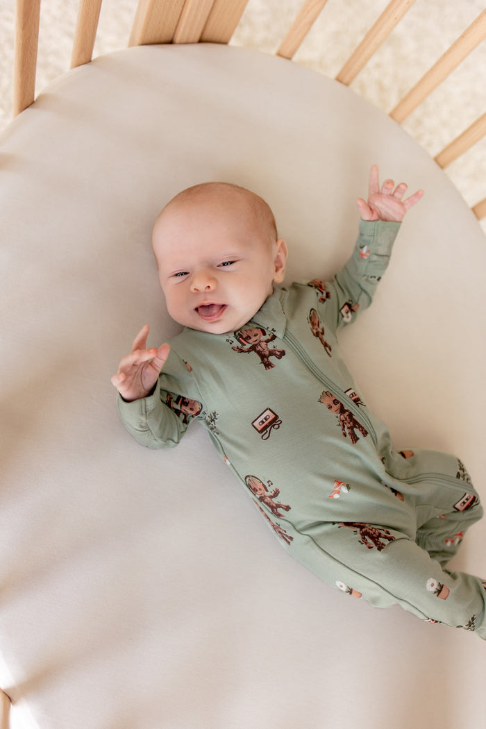 Baby lying in an oval cot wearing Green Zippered Footie in Groot print on a beige bassinet sheet