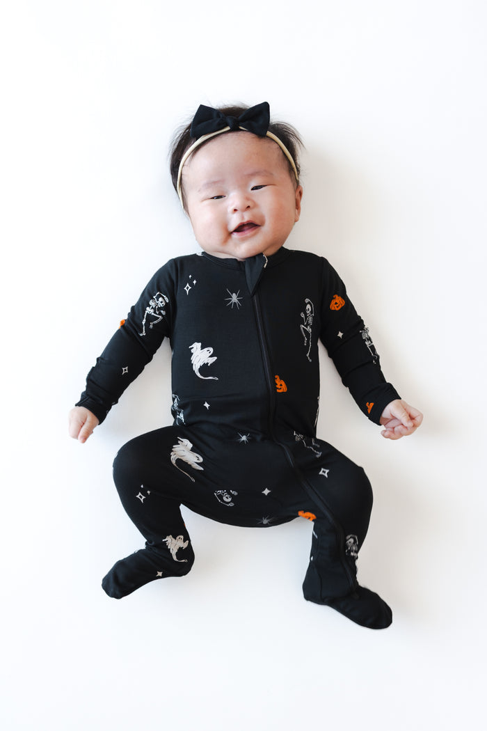 Baby wearing Zippered Footie in Halloween Friends print and a black bow on a white background