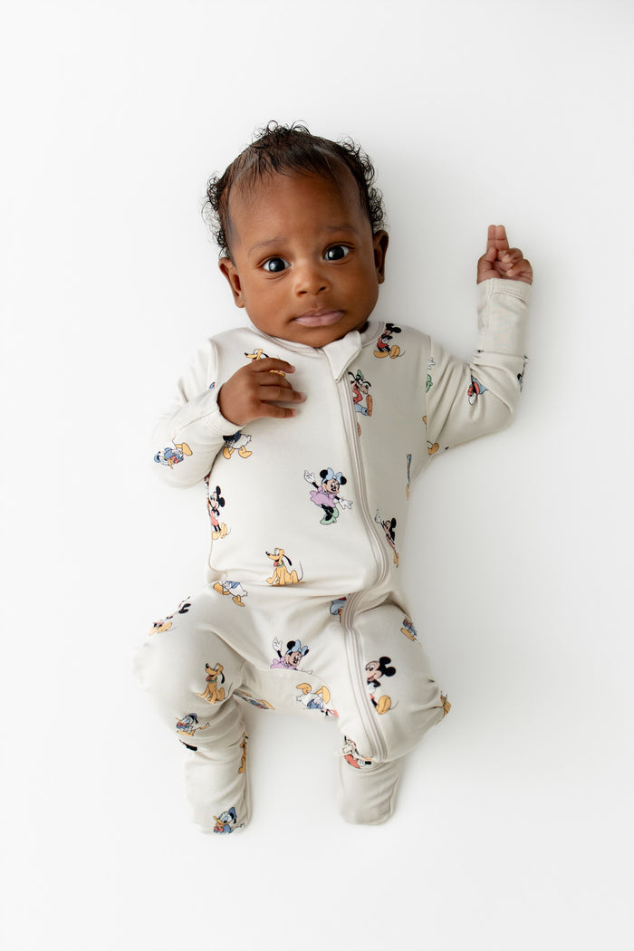 Baby wearing a cream zippered footie with colorful mickey cartoon character prints on a white background