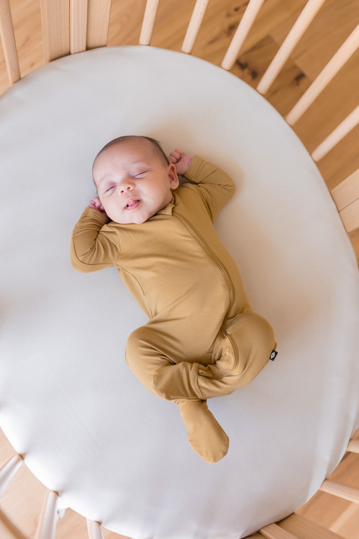 A sleeping newborn baby wearing a mustard yellow long-sleeve footed sleeper, lying on a white mattress inside a wooden crib.