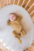 A sleeping newborn baby wearing a mustard yellow long-sleeve footed sleeper, lying on a white mattress inside a wooden crib.
