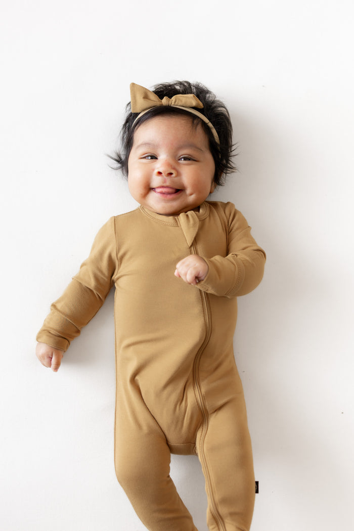 A smiling baby wearing a tan long-sleeve footed onesie and matching bow headband, lying on a white background.