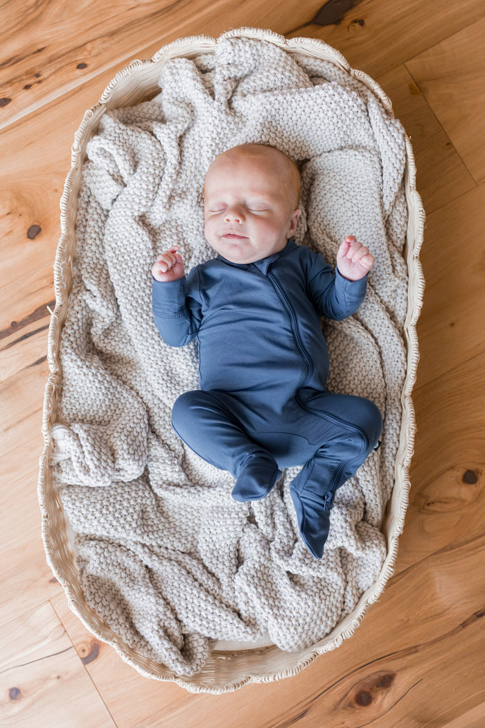 A sleeping newborn baby wearing a navy blue footed zipper onesie, lying on a textured cream knit blanket inside a woven oval bassinet on a wooden floor.