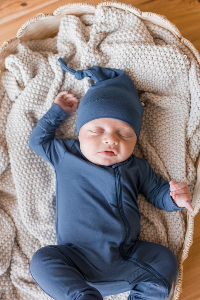 A sleeping newborn baby wearing a slate blue one-piece footie and matching hat, lying on a textured cream knit blanket inside a woven basket.