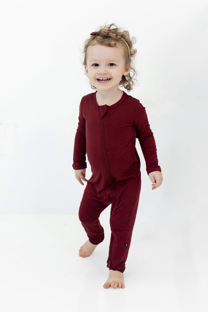Toddler standing wearing the Zippered Romper in Burgundy with matching tiny nylon bow