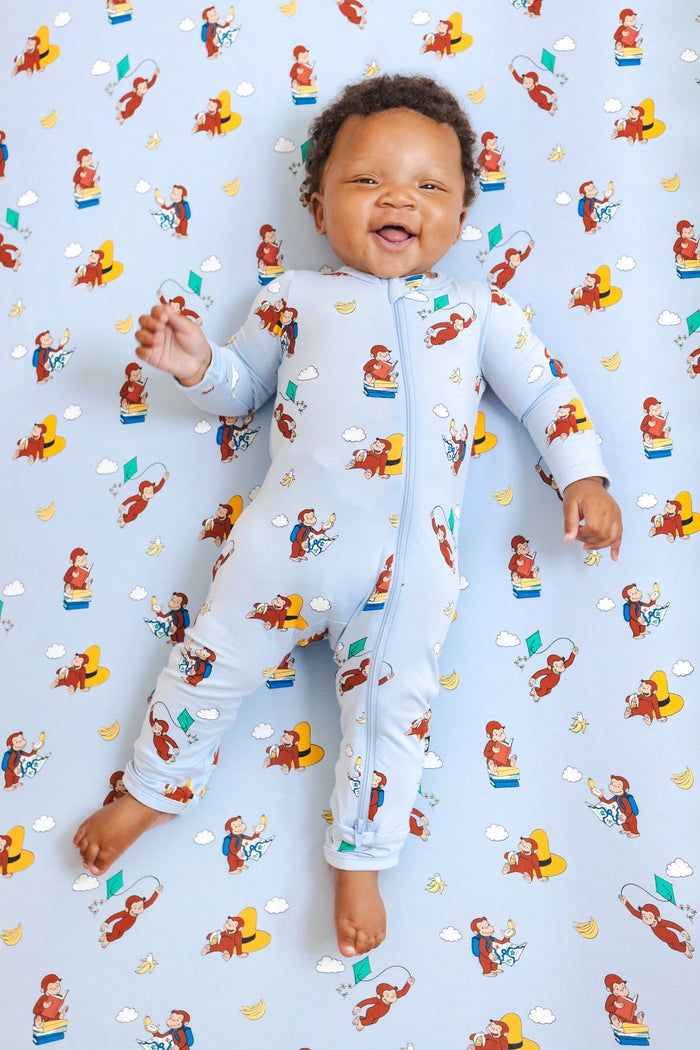 Baby smiling in Zippered Romper in Curious George print lying over a similar print crib sheet