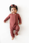 A baby lying on a white background wearing a dusty terracotta long-sleeve footless one-piece romper and a matching bow headband.