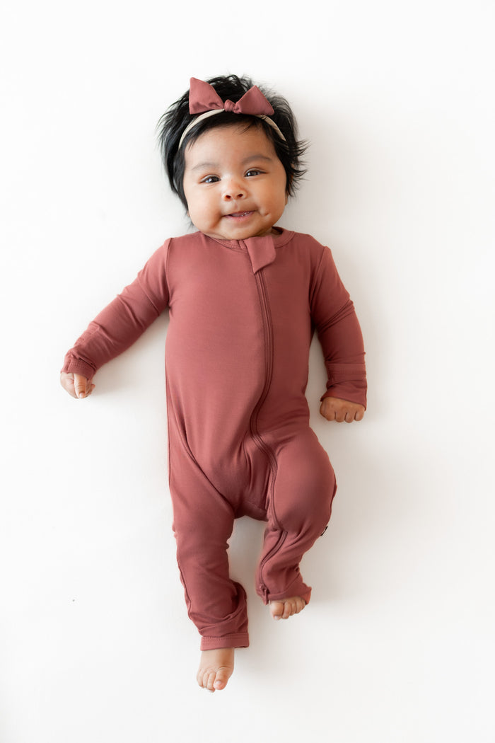 A baby lying on a white background wearing a dusty terracotta long-sleeve footless one-piece romper and a matching bow headband.
