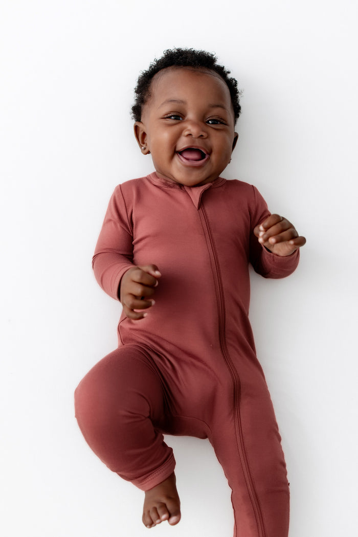 A smiling baby wearing a terracotta zip-up footless romper, lying on a clean white background.
