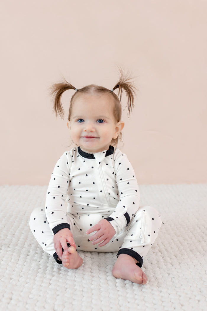 Child sitting in zippered romper with midnight polka dot print on beige background.