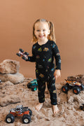 Girl holding toy truck wearing black Zippered Romper in Monster Truck print on sand and rocks with toy trucks